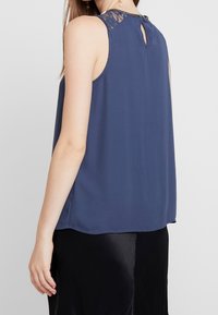 Haut bleu marine sans manches avec des accents en dentelle aux épaules, un détail fendu au dos et une texture lisse et fluide.