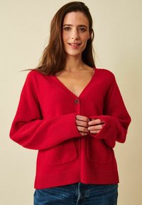 Kvinna med långt brunt hår, klädd i en röd kofta med knappar och blå jeans, stående mot en beige bakgrund och leende milt.