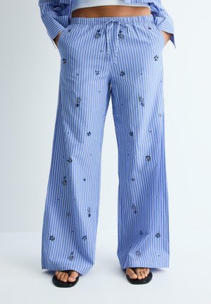 Pantalon large rayé bleu et blanc avec petite broderie florale, taille à cordon, porté avec des tongs noires et un top court.