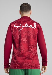 Burgunderfarbenes langärmliges Trikot mit einem dezenten geometrischen Muster, weißem arabischem Text "المغرب" auf der Rückseite, in taillierter Form und mit weicher Textur.