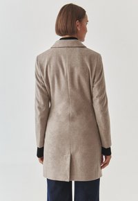 Abrigo de lana beige con un diseño estructurado, cuello con muesca, mangas largas y una abertura central en la parte posterior, combinado con acentos oscuros en los puños.