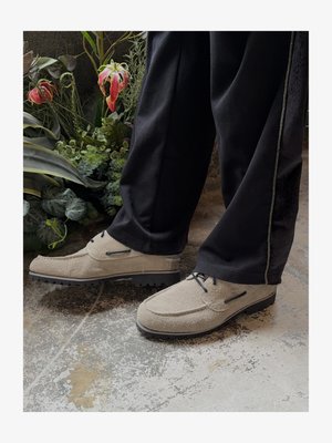 Φωτεινά μπεζ υφασμάτινα παπούτσια με κορδόνια, φορεμένα με μαύρο παντελόνι, στέκονται σε παλιό, υφασμάτινο δάπεδο δίπλα σε πράσινα και κόκκινα φυτά.