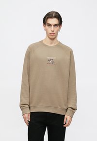 Καφέ φούτερ με στρογγυλή λαιμόκοψη, ριμπ μανσέτες και ζώνη μέσης. Διαθέτει κεντημένο λογότυπο στο στήθος και μανίκια ράγκλαν. Φορεμένο με μαύρο παντελόνι.