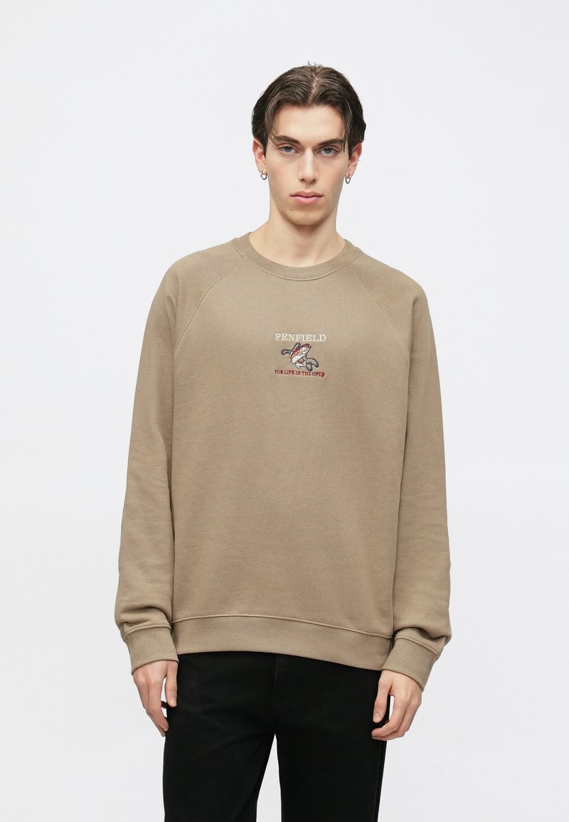 Καφέ φούτερ με στρογγυλή λαιμόκοψη, ριμπ μανσέτες και ζώνη μέσης. Διαθέτει κεντημένο λογότυπο στο στήθος και μανίκια ράγκλαν. Φορεμένο με μαύρο παντελόνι.