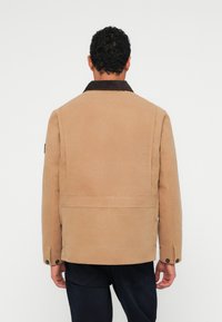 Brun canvasjakke med fløjlskrave. Har en lige pasform og knapmanschetter. Bagerste visning viser enkle sømme og minimal detaljering.