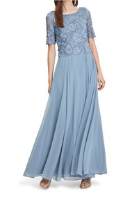 Kvinna klädd i en flödande dammig blå klänning med spetsliv, korta ärmar och en lång plisserad kjol, tillsammans med sandaler med remmar.