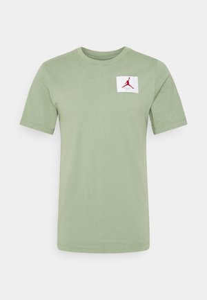 Jasnozielona bawełniana koszulka z krótkimi rękawami i okrągłym dekoltem, z białą łatą z czerwoną grafiką logo po lewej stronie klatki piersiowej.