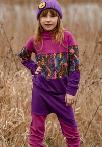 Fioletowy i różowy strój z wzorzystym panelem ozdobionym kolorowymi liśćmi. Do tego fioletowa dziergana czapka z naszywką w kształcie uśmiechniętej buźki.