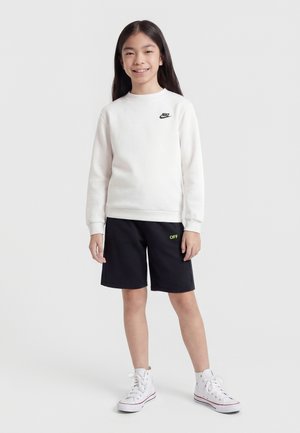 Νεαρό άτομο που στέκεται και χαμογελά, φορώντας λευκή φούτερ Nike, μαύρη βερμούδα με κίτρινο κείμενο "OFF" και λευκά ψηλά αθλητικά παπούτσια.