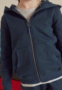 Hanorac cu fermoar închis în față, de culoare albastru închis, cu textură moale, buzunare frontale și un tricou gri dedesubt. Dispune de glugă cu șnur și fermoar argintiu.