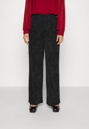 Persoon draagt zwarte broek met wijde pijpen met een zilverkleurig gespikkeld patroon, gecombineerd met zwarte schoenen en een rode blouse met lange mouwen.