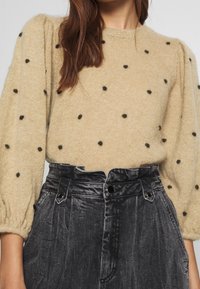 Pull en tricot beige avec des pois noirs texturés et des manches bouffantes, associé à un pantalon en denim gris foncé à taille haute avec une fermeture à boutons.