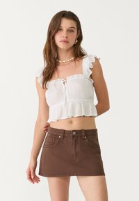 Biely volánkový crop top má uväzovanie vpredu, skombinovaný s hnedou džínsovou mini sukňou s prednými vreckami a kovovým zapínaním.