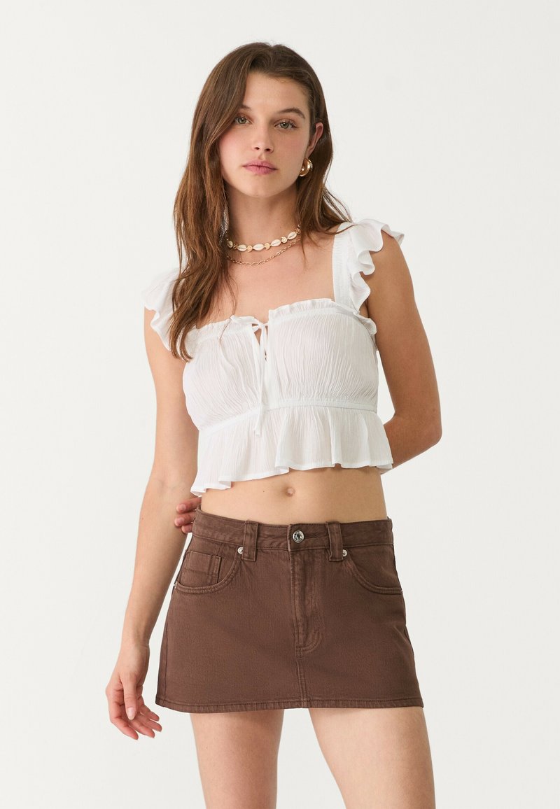 Biely volánkový crop top má uväzovanie vpredu, skombinovaný s hnedou džínsovou mini sukňou s prednými vreckami a kovovým zapínaním.