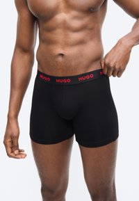 Άνδρας που φοράει μαύρα μποξεράκια με κόκκινο λογότυπο "HUGO" στη μέση, προσαρμόζοντας τη μέση στη δεξιά πλευρά του.