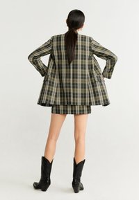 Ruutukuviollinen bleiseri ja minihame setti hillityissä vihreän ja mustan sävyissä. Bleiserissä on rento istuvuus ja kaksi taskua. Malli käyttää mustia cowboy-saappaita.