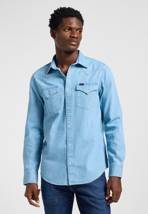 Mann trägt hellblaues Jeanshemd mit Brusttaschen, blickt leicht nach rechts vor einem einfachen Hintergrund.