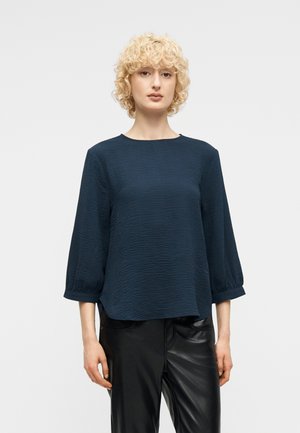 Kobieta o krótkich, kręconych blond włosach nosi teksturowaną, ciemnoniebieską bluzkę z długim rękawem i czarne skórzane spodnie na jednolitym tle.