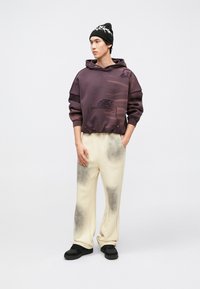 Sweatshirt oversize roxa com detalhes desgastados, combinada com calças de ganga cremes com manchas cinzentas e ténis pretos. Gorro preto na cabeça.