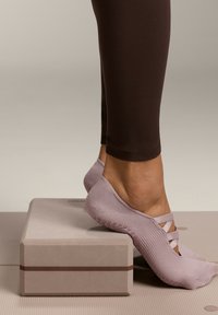 Sosete de grip roz pal cu o talpă texturată, dotate cu bretele încrucișate, așezate pe un bloc de yoga gri pe un fundal bej.