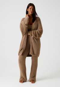 Cardigan beige fuzzy con grandi bottoni, abbinato a pantaloni aderenti coordinati. Tessuto morbido e vestibilità rilassata. Modella in piedi scalza contro uno sfondo neutro.