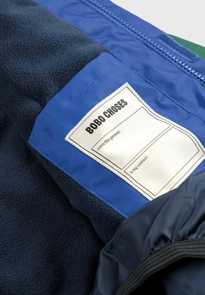 Veste en polar avec un patch en tissu bleu et une étiquette, corps marine, fermeture éclair et poignets élastiques, mettant en valeur la texture et le contraste.