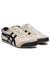 Beige leather sneakers with a navy blue design, suede accents, flat laces, and a textured black rubber sole. Classic athletic style.