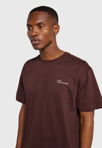 Man med kort hår och örhänge som bär en mörkbrun T-shirt med vit text "HALO 196819721981" på bröstet, mot en enfärgad bakgrund.