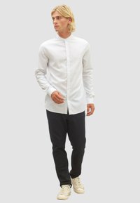 Weißes Langarmshirt mit Stehkragen, kombiniert mit dunklen Slim-Fit-Hosen und beigen Sneakers, stehend vor einem neutralen Hintergrund.