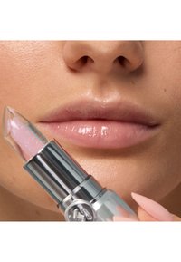 Close-up of glossy natural lips with a hand holding a clear, glittery lipstick near the mouth.