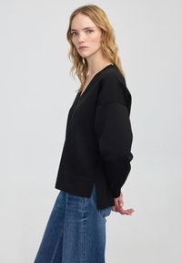 Zwarte, langermige trui met een V-hals. Heeft een relaxte pasvorm, zijsplitten en een gestructureerde stof, gecombineerd met blauwe denim jeans.