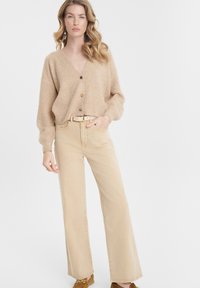 Cardigan lavorato a maglia beige con scollo a V e bottoni di legno, abbinato a jeans chiari beige a vita alta e pantaloni larghi con scarpe marroni slip-on.