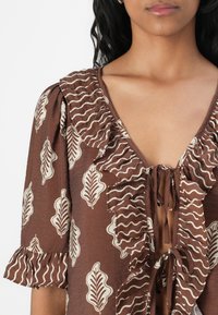 Blusa marrone con scollatura a V e dettagli intrecciati, bordi arricciati e un motivo a foglia e linee ondulate in crema. Maniche corte con rifinitura arricciata.