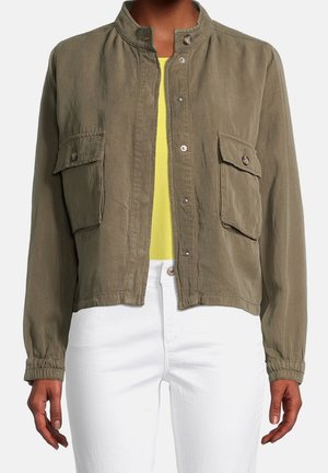 Giacca verde oliva con bottoni e due tasche con pattina davanti, indossata sopra un top giallo e abbinata a pantaloni bianchi.