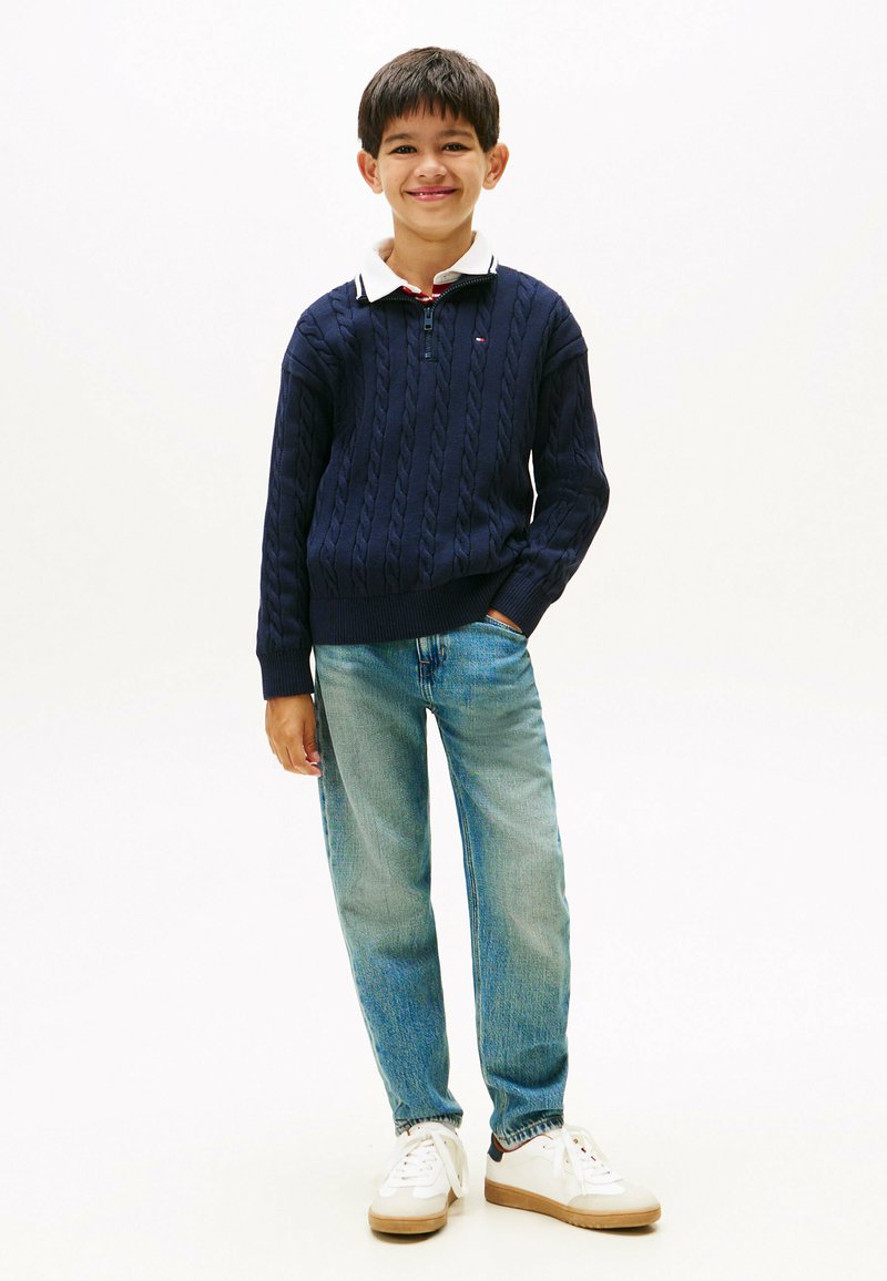 Jūras spēku krāsas adījuma džemperis ar rāvējslēdzēju, zem tā balta apkakles krekla, gaiši zili džinsi un baltas kedas ar brūniem zoliem.