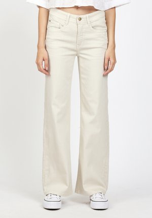 Frau trägt off-white weite Jeans und weiße Sneakers, steht aufrecht vor einem schlichten hellen Hintergrund.