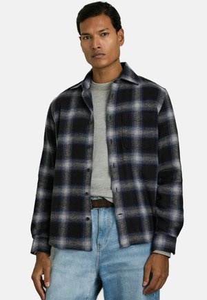 Cămașă din flanel în carouri, cu culori albastre, negre și albe, cu nasturi în față, guler deschis și un buzunar pe piept, purtată peste un tricou gri.