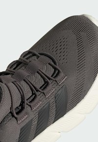 Gray athletic shoe featuring a textured mesh upper, flat laces, and three black accent stripes on the side. White rubber sole.