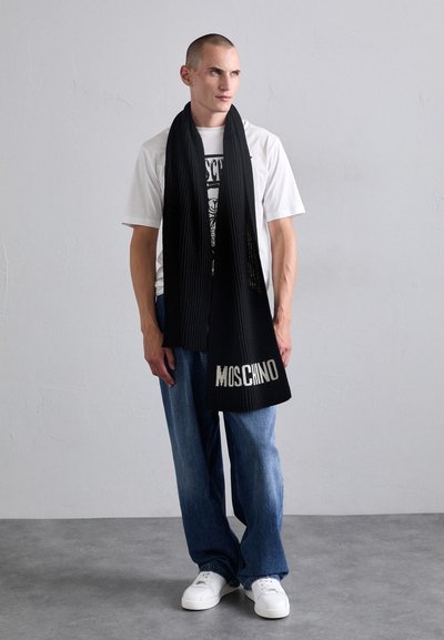 Melna ribota šalle ar "MOSCHINO" logo apakšā, kombinēta ar baltu T-kreklu un gaiši ziliem džinsiem. Audums ir mīksts un adīts.