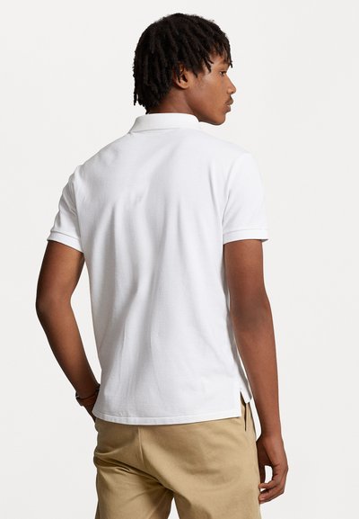 Mann mit kurzen Dreadlocks, der ein weißes kurzärmeliges Poloshirt und beige Hose trägt, steht mit dem Rücken und seitlich gedrehtem Kopf.