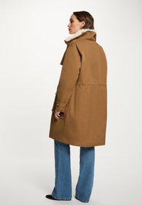 Cappotto oversized marrone con colletto alto e foderato in pelliccia sintetica, caratterizzato da una vita fasciante e maniche con due bottoni, abbinato a jeans a gamba larga.