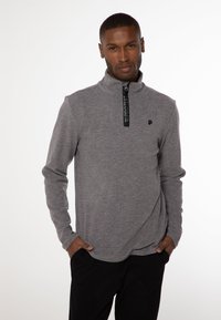 Grå zip-up pullover lavet af et tekstureret stof, med en sort lynlås med branding og et lille logo på brystet. Lange ærmer.