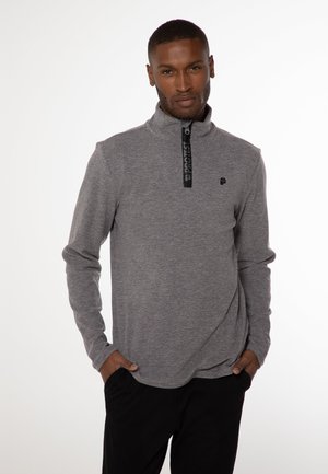 Grå zip-up pullover lavet af et tekstureret stof, med en sort lynlås med branding og et lille logo på brystet. Lange ærmer.
