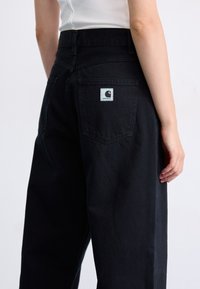 Zadný pohľad na osobu v čiernych vysokých jeansoch Carhartt a bielom tielku na hladkom svetlom pozadí.