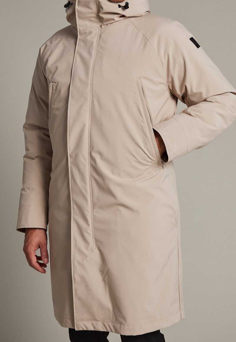 Beige lång kappa med dragkedja fram, justerbar huva, sidofickor och en slät, matt tygtextur. Har en distinkt svart logotyp.