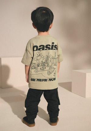 Gyerek bézs színű pólót visel, rajta az "oasis" felirat és egy utcai jelenet grafikája, fekete bőrnadrággal és barna talpú cipővel, háttal állva.