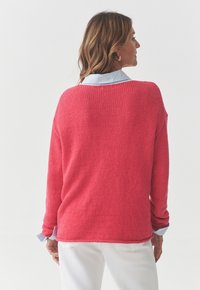 Maglione rosa a maglia con vestibilità ampia, caratterizzato da una superficie texturizzata e un colletto azzurro sottostante a strati. Sono visibili dei pantaloni bianchi.