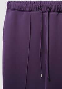 Lila Sporthose mit elastischem Bund, Kordelzug und Seitentaschen. Glatte Textur mit einem dezenten Glanz auf dem Stoff.