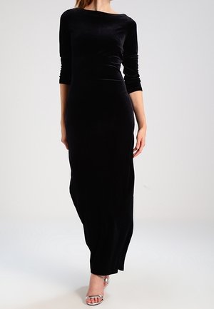 Kobieta ubrana w długą czarną welwetową sukienkę z rękawami do połowy łokcia i srebrne, otwarte szpilki na wysokim obcasie, stojąca na jednolitym tle.