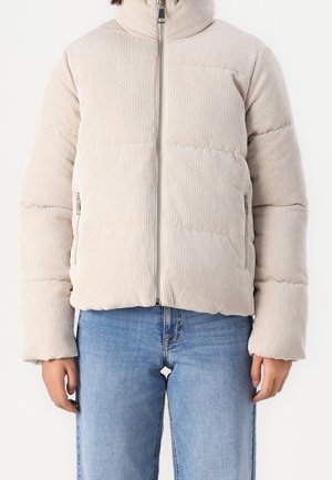 Personne portant une veste matelassée en velours côtelé beige avec fermeture éclair devant et un jean bleu clair, debout devant un fond uni.